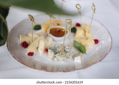 Four Different Types Of Cheere On A Handmade Plate. Bamboo Toothpicks In Cheese. Mustard Honey Sauce And Cranberries Around Dor Blue, Parmesan, Camembert And Cheddar Cheese. Outdoor Feast On Party