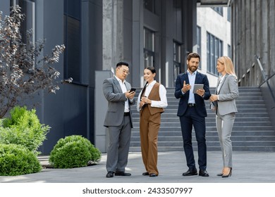 Four diverse business professionals engage in a casual meeting outdoors. They are using smartphones and tablets while discussing work-related matters, with a corporate building in the background. - Powered by Shutterstock