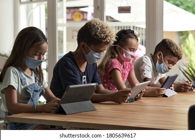 Four diverse kids wear facemasks sit at table use wireless gadgets ignoring each other prefer internet games and virtual communication. Alpha generation and modern technology overuse, phubbing concept
