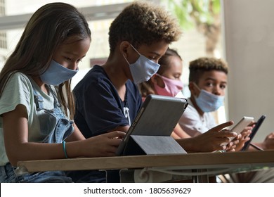 Four diverse kids wear facemasks sit at table use wireless gadgets ignoring each other prefer internet games and virtual communication. Alpha generation and modern technology overuse, phubbing concept - Powered by Shutterstock