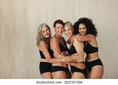 Four Diverse Women Of All Ages Embracing Their Bodies. Group Of Smiling Women Wearing Black Underwear And Embracing Each Other Against A Studio Background.