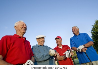 Four elderly men are standing together on a golf course. They are holding their clubs, smiling, and looking away from the camera.  Horizontally framed shot.