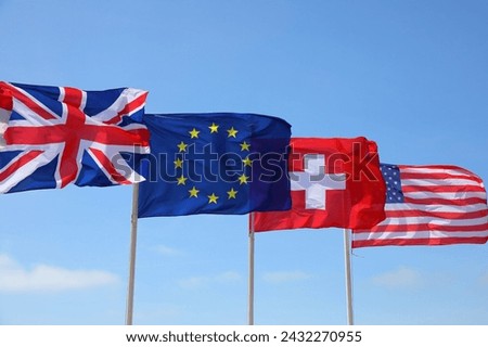 Four flags waving in the breeze on four flagpoles against a blue sky on a sunny day.
