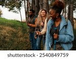 Four friends hiking in a forest, wearing casual outdoor clothing and carrying trekking poles. They are walking in a line along a grassy path surrounded by trees.