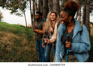 Four friends hiking in a forest, wearing casual outdoor clothing and carrying trekking poles. They are walking in a line along a grassy path surrounded by trees. - Powered by Shutterstock