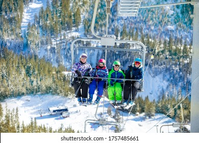 Four Friends Kids Sit High Together On Chairlift Lifting On The Mountain With Green Forest Below