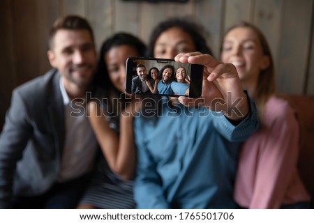 Four friends making selfie, photo on smart phone screen view, focus on hand holds device. Modern tech usage, have fun party event hanging out, taking memory pic, multi ethnic people friendship concept