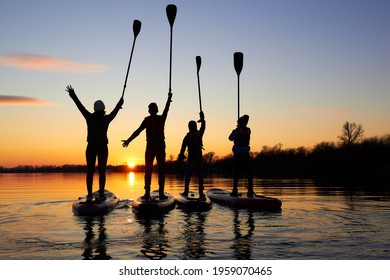 Four friends on stand up paddle board (SUP) on a flat quiet winter river at sunset raising his paddles up