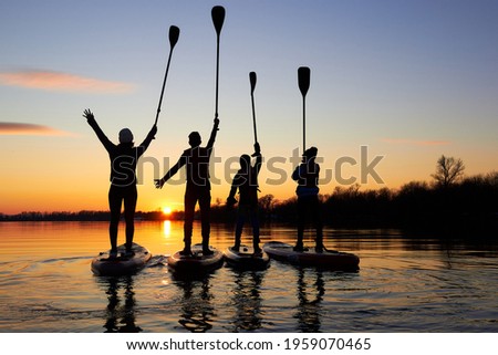 Four friends on stand up paddle board (SUP) on a flat quiet winter river at sunset raising his paddles up