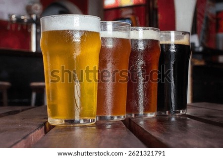 Four glasses filled with different types of craft beer on a rustic wooden table inside a bar. Warm ambient lighting, no people, cozy pub atmosphere, beer variety on display.