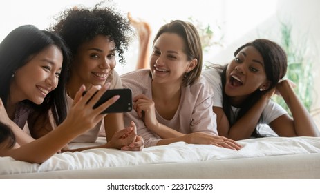 Four gorgeous multiracial girls friends enjoy time together. lying in bed. hold smart phone. Womans using new application, have fun on-line hanging out, awaken after sleepover. Friendship funny - Powered by Shutterstock