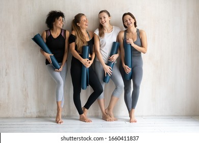 Four gorgeous slim multicultural young women wearing stylish comfy sporty wear holding yoga mats ready for workout at gym posing against wall background. Girls keeping fit and healthy wellness concept