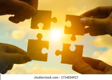 Four Hands Of Businessmen Assemble Pieces Of A Puzzle Into A Single Whole Against The Backdrop Of The Sunset, Business Concept Idea, Partnership, Teamwork, Cooperation, Creative