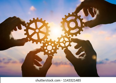 Four hands of businessmen collect gear from the gears of the details of puzzles. against the backdrop of dramatic sunlight. The concept of a business idea. Teamwork, strategy, cooperation, creativity