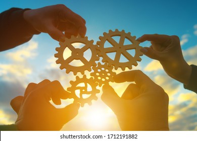 Four Hands Of Businessmen Collect Gear From The Gears Of The Details Of Puzzles. Against The Background Of Sunlight. The Concept Of A Business Idea. Teamwork. Strategy. Collaboration, Partnership