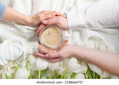 four hands hold one of heart-shaped cofee cup, white tulips