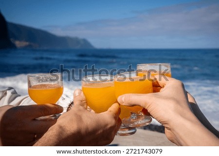 Four hands raise small goblets of orange poncha as waves meet volcanic cliffs near Porto Moniz, Madeira, Portugal. Warm daylight, crisp glasses, soft distant coast.