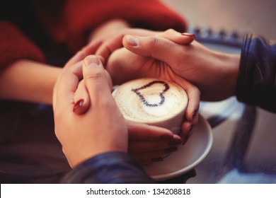 Four hands wrapped around a cup of coffee with heart drawing