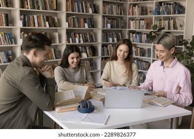 Four happy attractive multi racial students learning subject using laptop, watch educational on-line video, studying together seated at desk gather in college library. Education, modern tech, teamwork