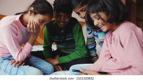 Four Happy South Asian Kids Sitting On The Ground And Looking At The Smartphone