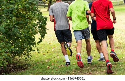 Four High School Cross Country Runners Training In A Park Running Around A Large Bush In A Group.