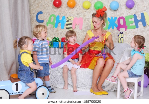 Four kids look\
at entertainer inflating balloon by pump at children party.\
Inscription Happy Birthday on\
wall.