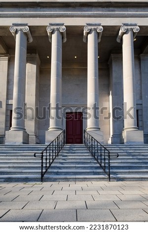 Four large round concrete columns at the top of marble steps with black iron rails to a legal building. The government building has a tall red door.  The view is from the lower corner of the steps.