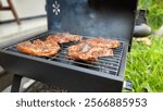 Four marinated and glossy lamb chops being grilled on a compact, black, portable charcoal barbecue, with a light haze of white smokes emanates from the grill at a a lush green grass backyard garden.