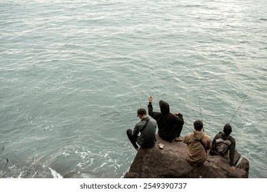 Four men sit on a rocky shore, fishing peacefully as the ocean waves gently crash below. The scene captures a moment of camaraderie, patience, and harmony with nature. - Powered by Shutterstock