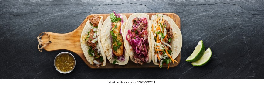 four mexican street tacos with fish barbacoa and carnitas shot in panoramic composition on top of serving wooden board