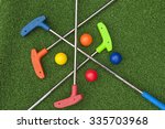 Four mini golf putters and balls of assorted colors laying criss crossed on artificial grass