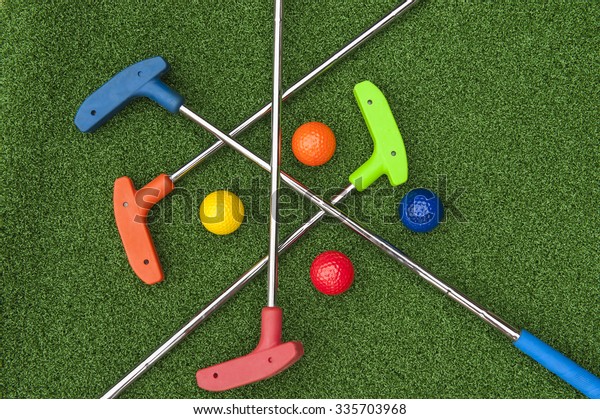 Four mini golf putters and balls of\
assorted colors laying criss crossed on artificial\
grass