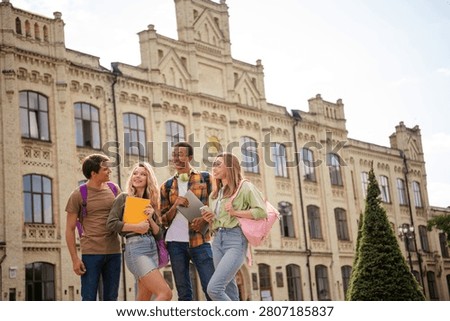 Four multicultural college students chat and laugh while carrying backpacks, notebooks and a tablet in front of a grand historic university building, conveying friendship, learning and campus life.