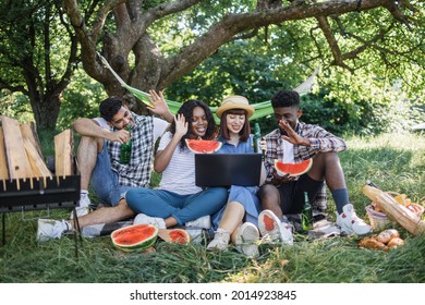 Four Multiracial Friends Smiling And Waving During Video Call On Modern Laptop While Relaxing On Nature. Young People With Alcohol And Food Having Picnic At Garden.