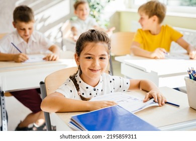 Four Nice Pupils Are Listening Carefully To Their Tutor. Primary School Kids Sitting On Desks And Working In Classroom. Education And School Concept.