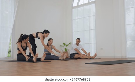 Four people in a bright, modern studio practicing seated forward bend poses with an instructor providing assistance. Group yoga class featuring diverse participants. - Powered by Shutterstock