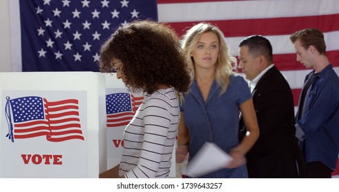 Four people of different demographics, young Hispanic woman in front, filling in ballots and casting votes in booths at polling station, US flag on wall at back