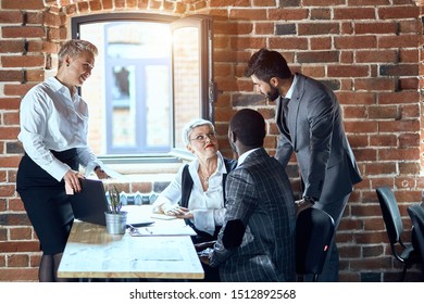 Four people dressed in stylish office clothes discuss task while sit at table. Window open. - Shutterstock ID 1512892568