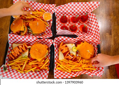 Four Plates Of Junk Food On The Table. Close Up Of Young Girls Holding Fries. Young Girls Hand Models Eating. Cheeseburgers And Fries On A Tray. Spicy Chicken Tender. American Cuisine At Restaurant.