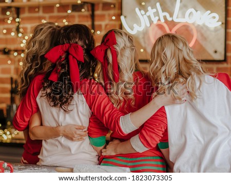 Four pretty women with curly hair and red Christmas bows in winter pyjamas posing in the kitchen. Christmas background. View from the back.