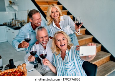 Four Senior Friends Having Dinner At Home. Two Handsome Mature Men And Two Attractive Women Spending Time Together Indoors. Group Of Old Friends Drinking Wine In Modern Apartment.