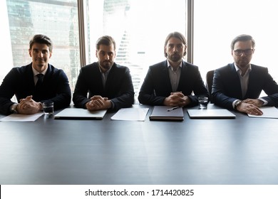 Four serious confident businessmen sitting at table in boardroom, looking at camera, focused hr managers listening to candidate, making decision, business partners gathering at corporate meeting - Powered by Shutterstock