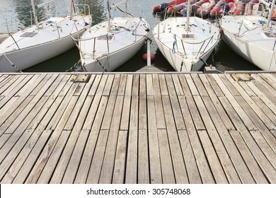 Four small learning sailing boats in a row in a wooden jetty. Big copy-space for placing text and some red pneumatic dinghies at background.