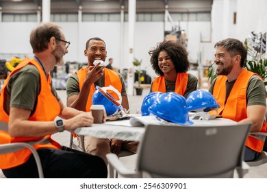 Four smiling multiracial engineers wearing orange safety vests are enjoying a coffee break at a round table in a factory, with blue safety helmets placed on the table