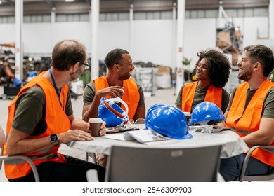 Four smiling multiracial engineers wearing safety vests are enjoying their coffee break, sitting around a table with blueprints and helmets in a factory - Powered by Shutterstock