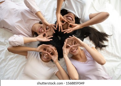 Four smiling young multi ethnic ladies lying on bed look at camera make binoculars, happy multiracial girls friends wear pyjamas having fun laugh together enjoy pajama hen party, top view portrait