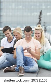 Four Students Sitting On The Ground. Two Are Watching At An Ipad And One Is Using His Mobile Phone. 