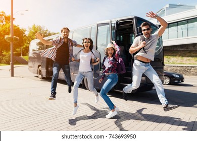 Четверо туристов прыгнули за позирование на фото. Они веселились. Они протянули руки и улыбнулись. На заднем плане находится современный черный автобус, на котором они будут ездить.