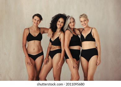 Four Women Of All Ages Celebrating Their Natural Bodies. Confident Women Smiling Cheerfully At The Camera While Wearing Black Underwear And Standing Together Against A Studio Background.