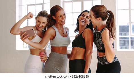 Four women in fitness wear standing together for a selfie after workout. Women laughing and showing muscles while standing for a selfie at a fitness studio. - Powered by Shutterstock